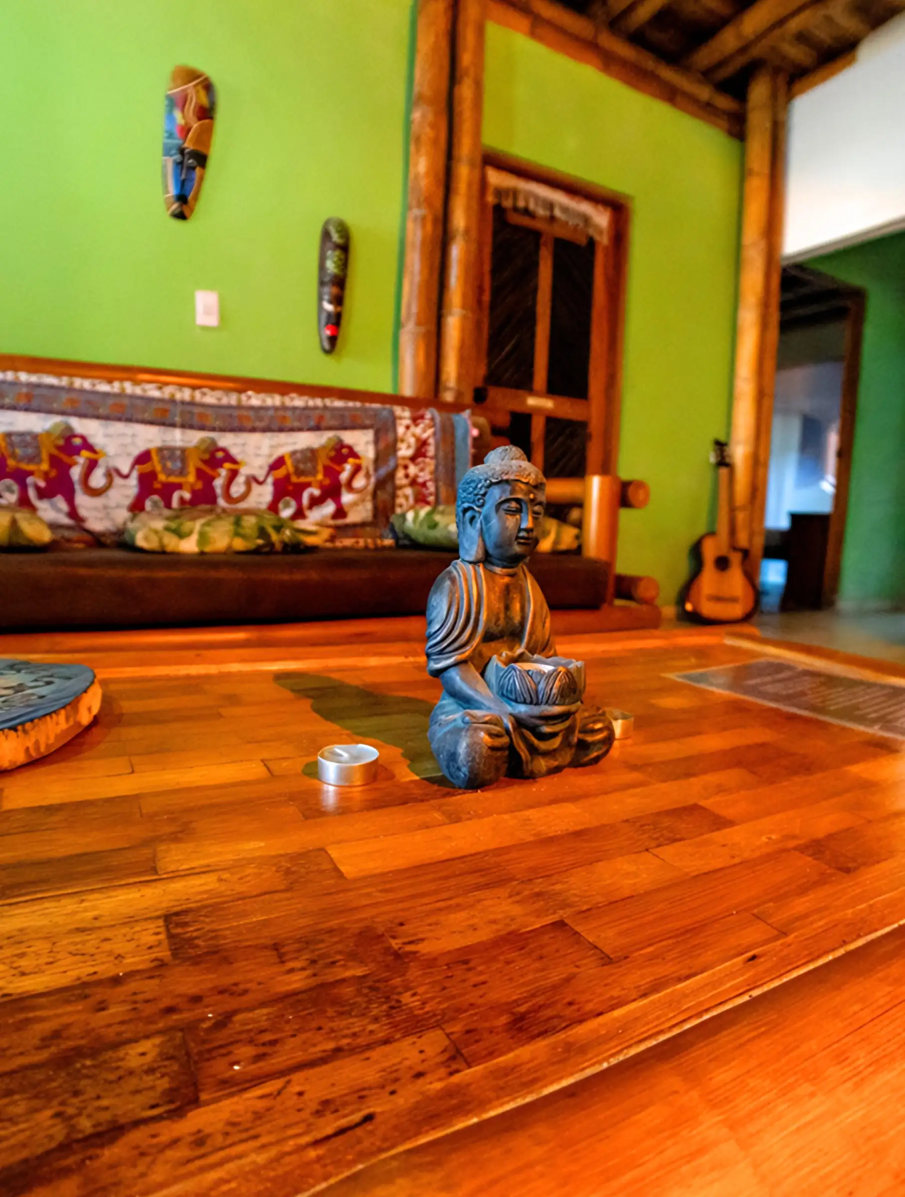 La sala común de paredes verde intenso: un Buda azul sobre el piso de madera, máscaras en la pared, un sofá con tela de elefantes y una guitarra.