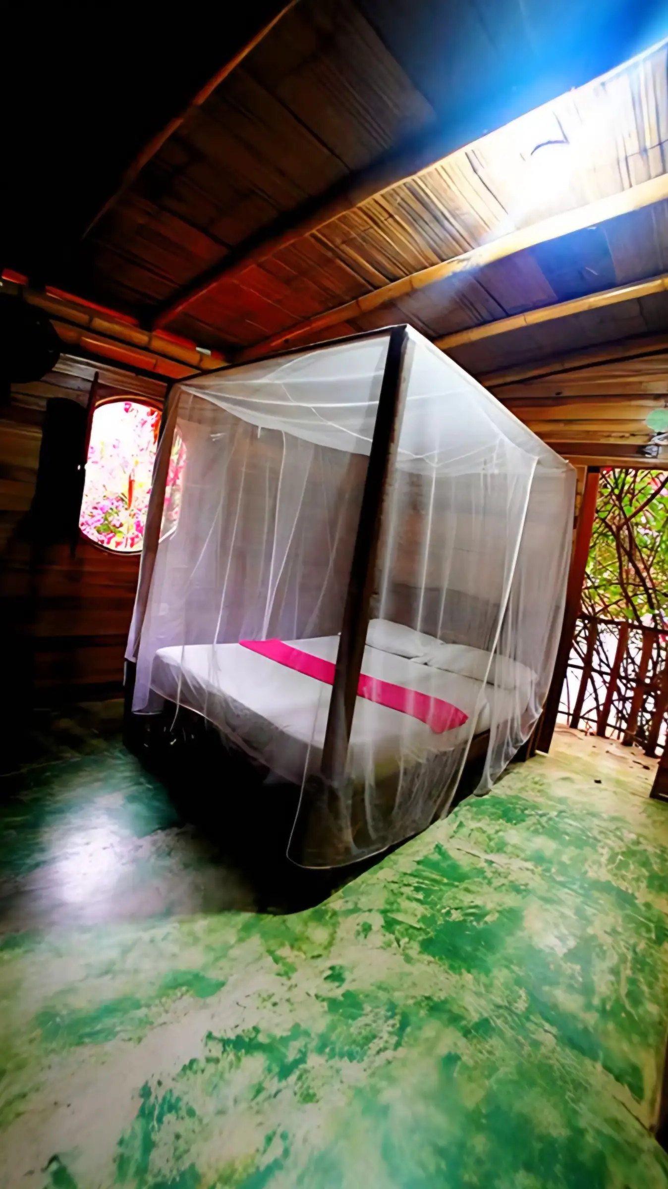 Habitación de bambú y madera con una cama con toldillo blanco, piso verde pintado a mano y una ventana redonda con flores afuera.