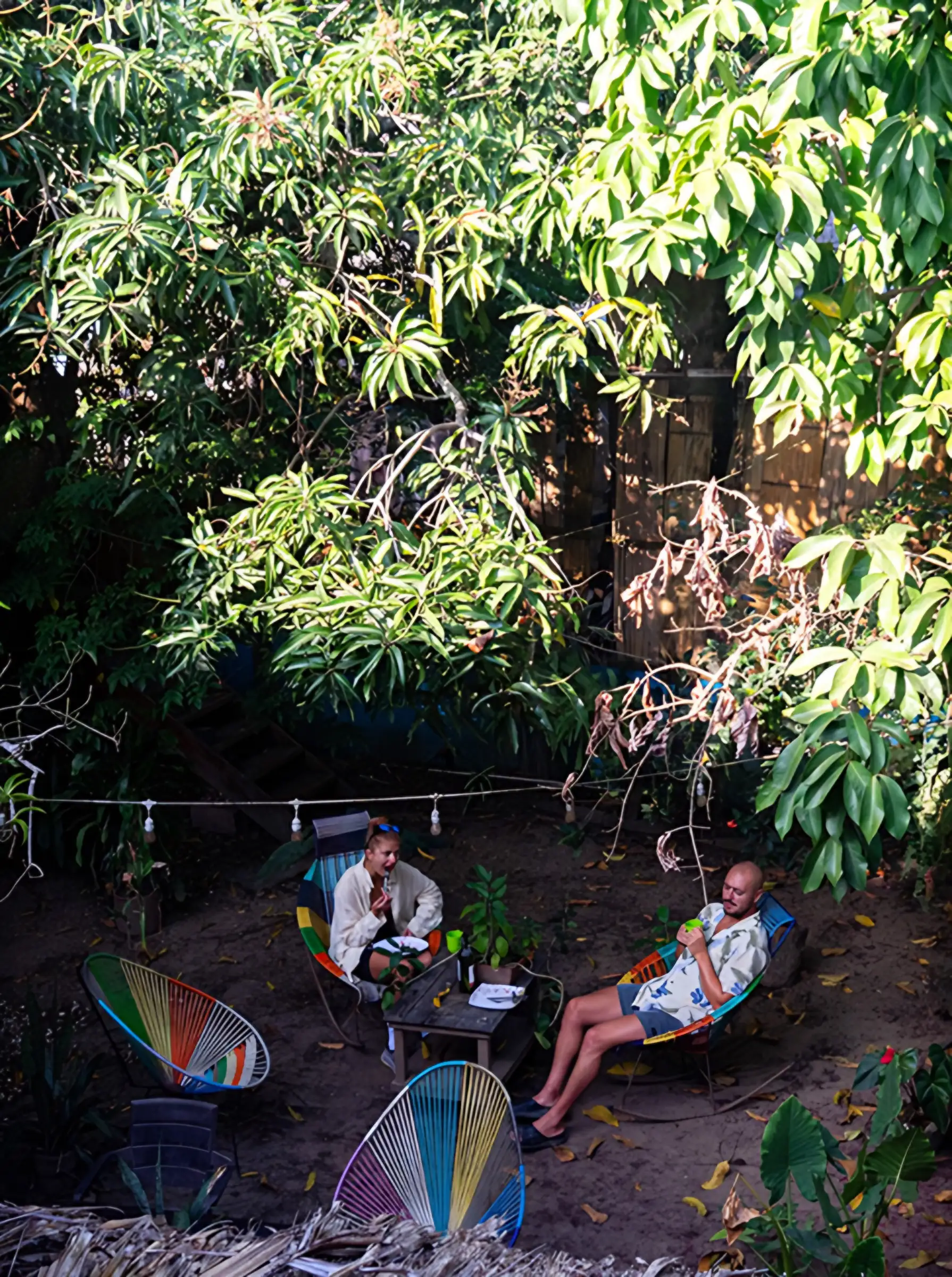 Dos huéspedes conversan en sillas Acapulco de colores bajo árboles frondosos, con luces colgantes en el jardín.