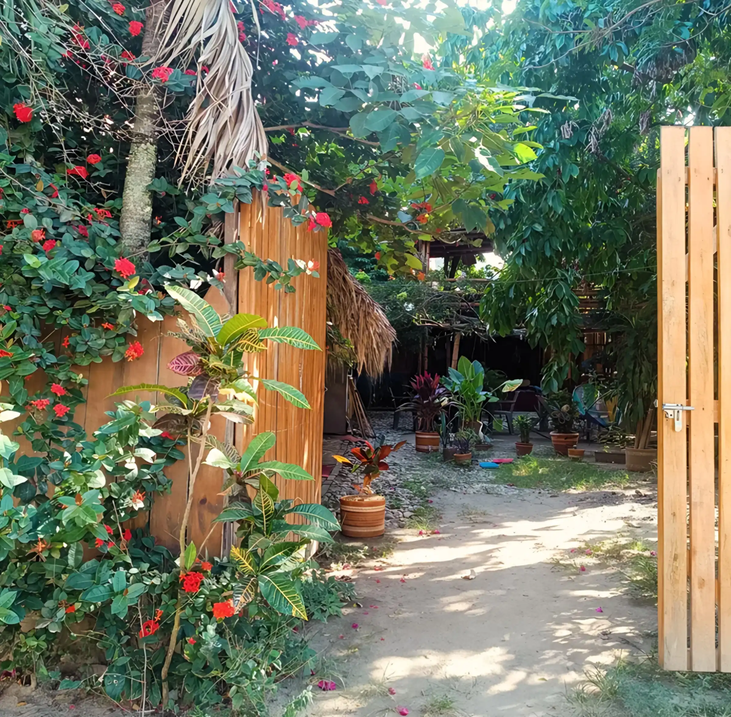 La puerta de madera del hostal abierta hacia el jardín: un camino de tierra entre plantas en materas, flores rojas de ixora y un kiosco con techo de palma al fondo.