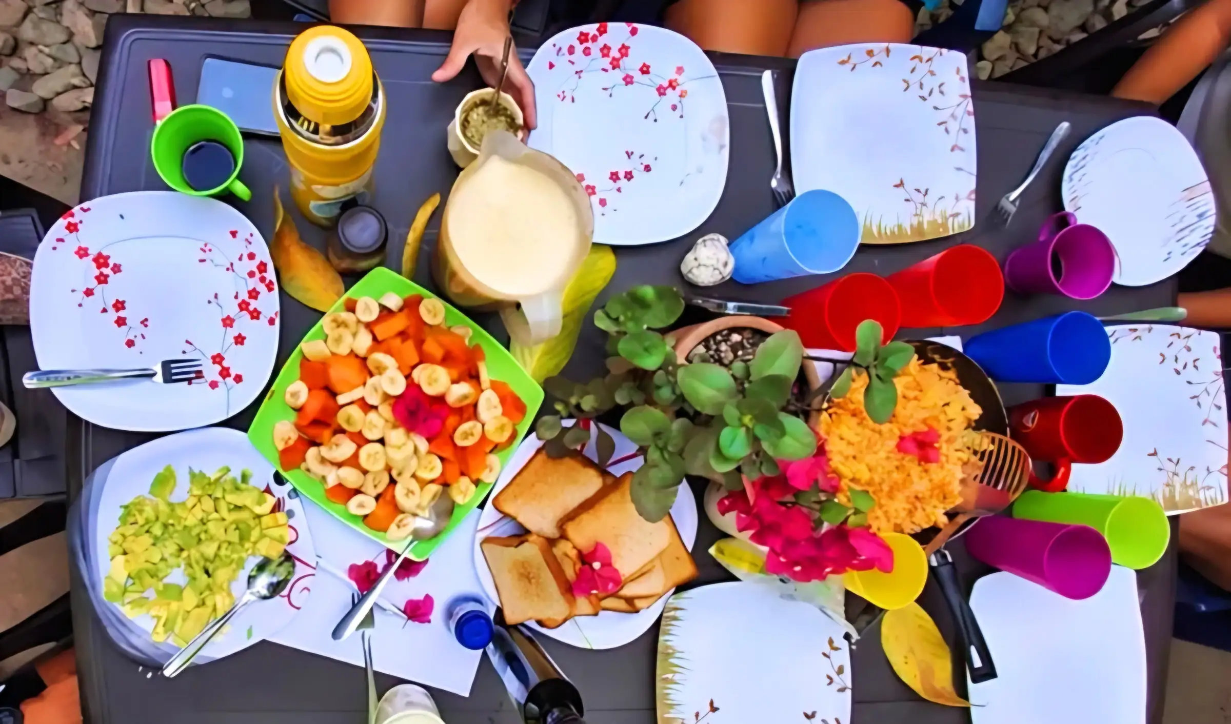 Una mesa vista desde arriba con fruta picada, huevos revueltos, tostadas, jugo y vasos de colores.