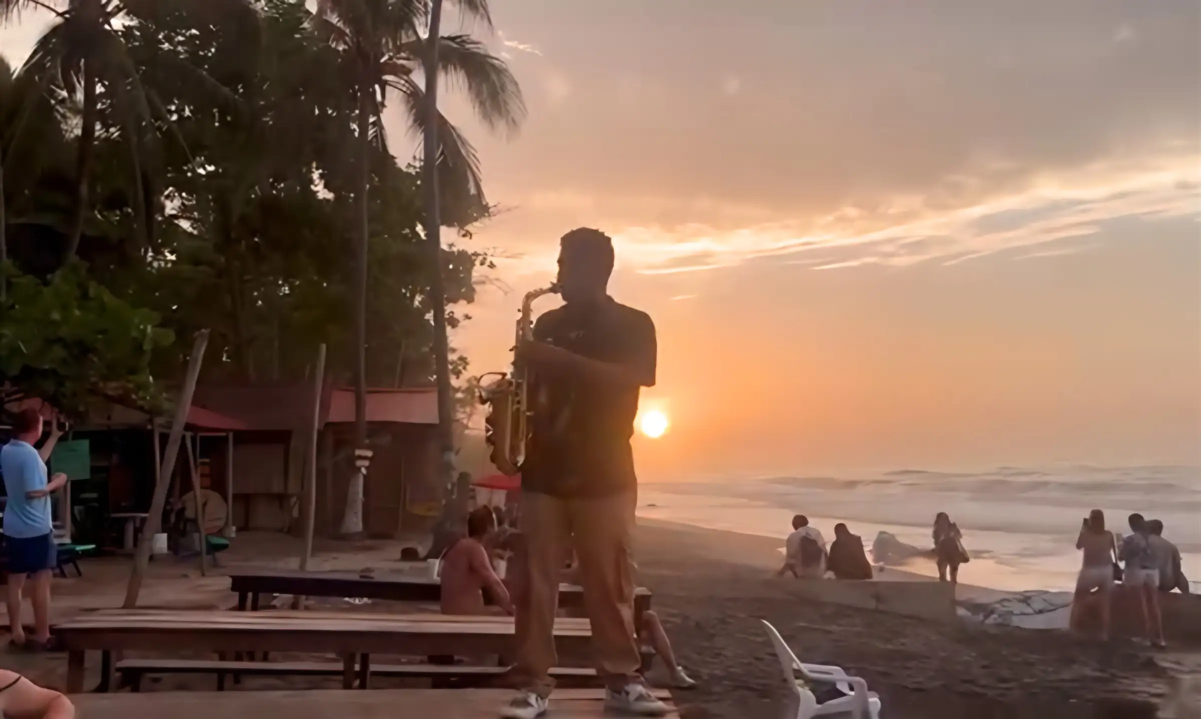 Un saxofonista toca en la playa al atardecer mientras la gente mira el mar.