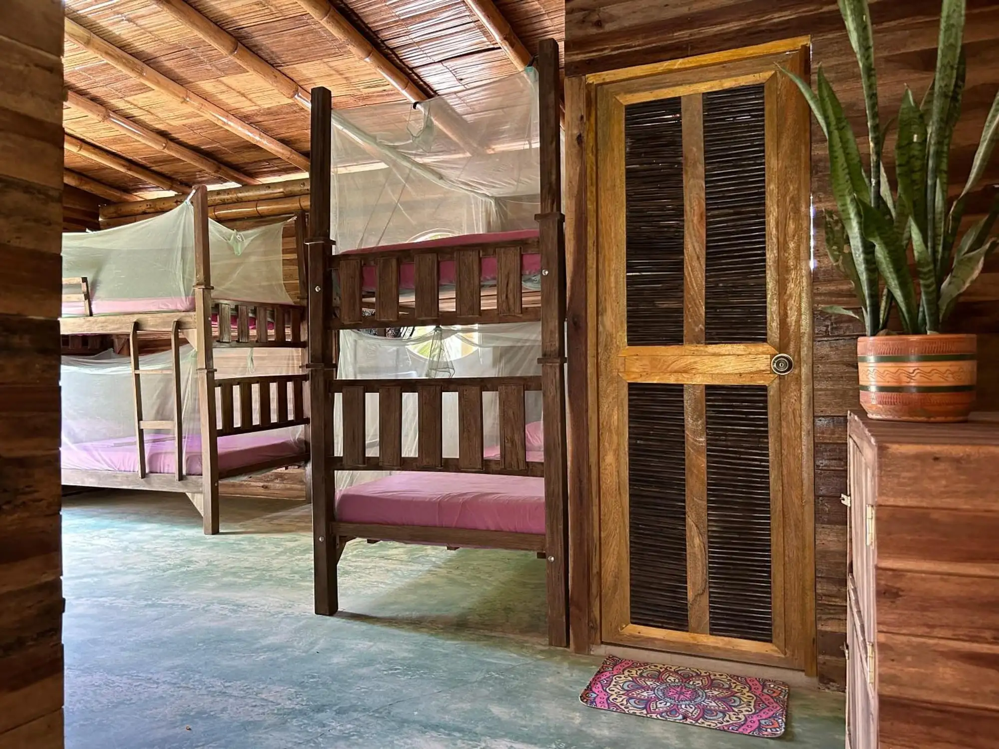 Un camarote de madera con toldillo junto a una puerta de celosía; al fondo, más camarotes del dormitorio.