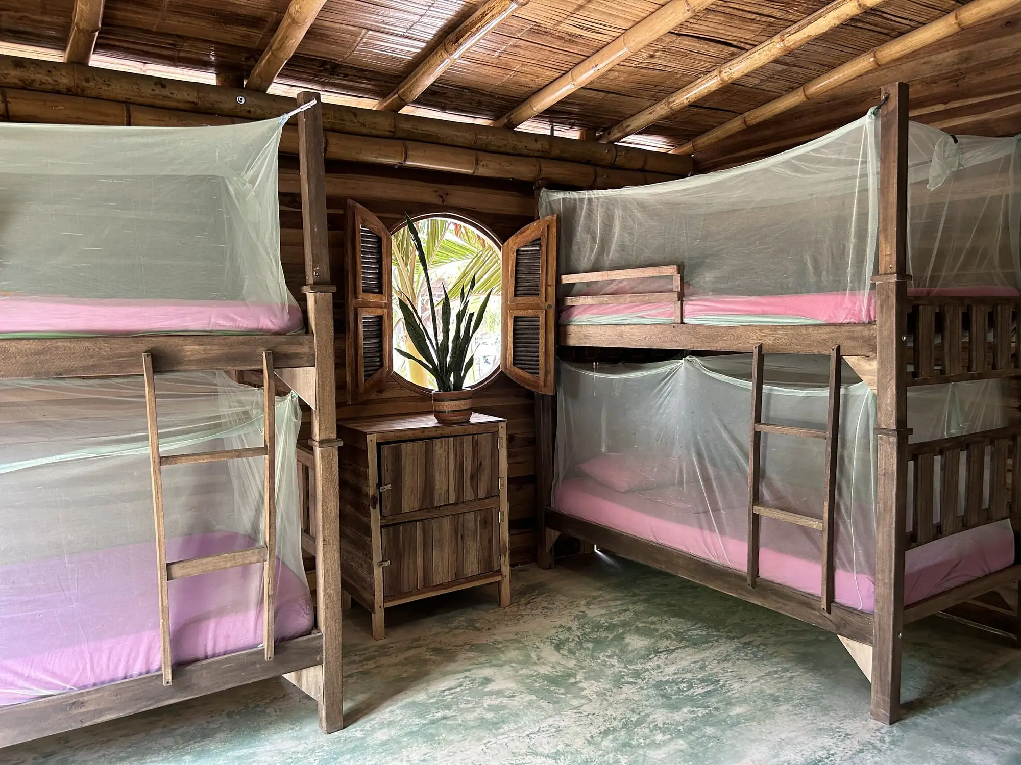 Dormitorio compartido de madera con techo de bambú: dos camarotes de madera con toldillos y una ventana redonda entre ellos.