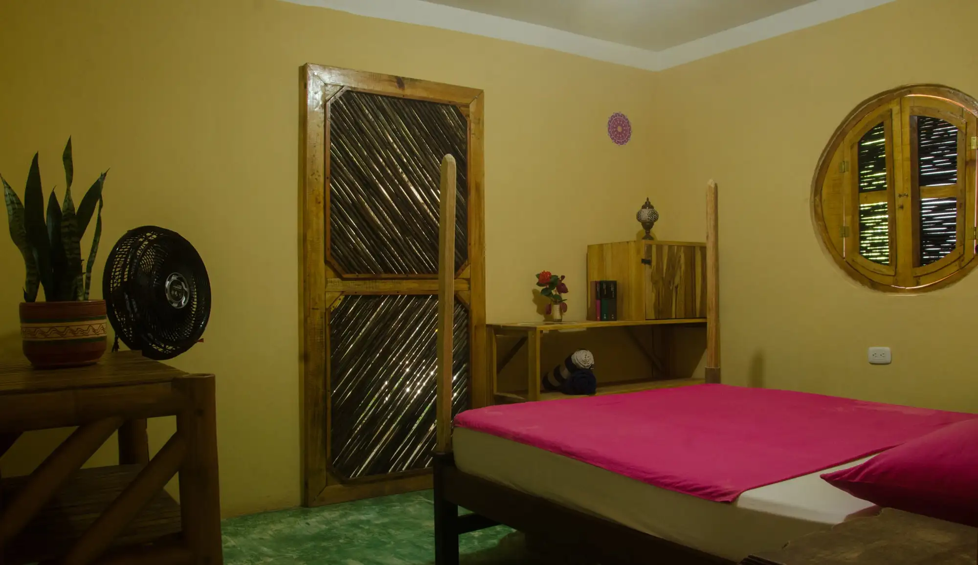 Habitación doble de paredes amarillas: cama doble con sábanas fucsia, puerta de madera con celosía, ventilador y ventana redonda.