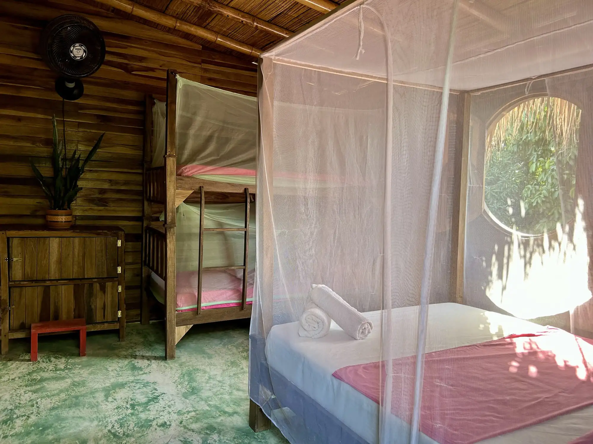 Habitación familiar de madera: cama doble bajo toldillo y un camarote al fondo, junto a una ventana redonda que da a la vegetación.