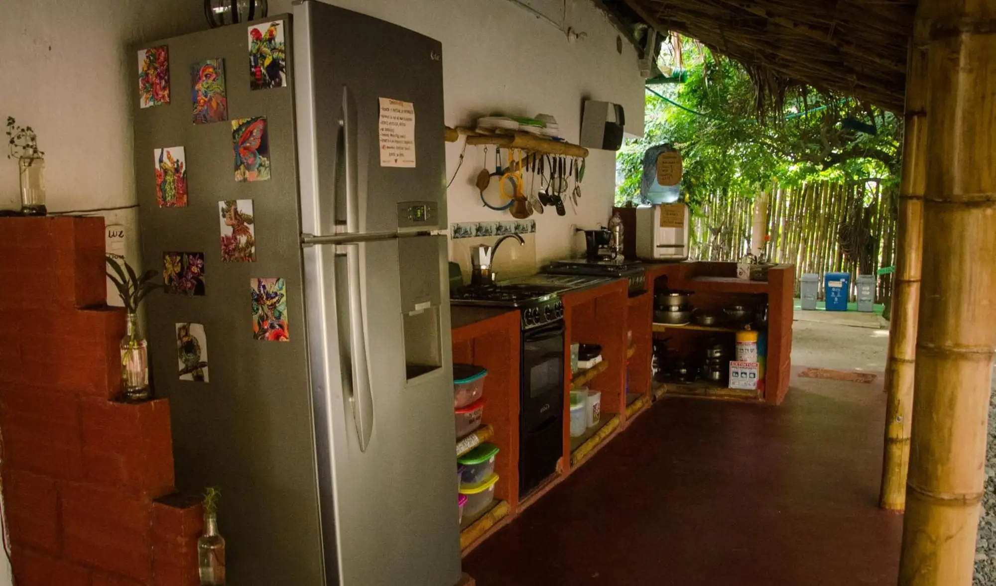 La cocina compartida: nevera con imanes, estufa, mesones de ladrillo y utensilios colgados, abierta hacia el jardín.