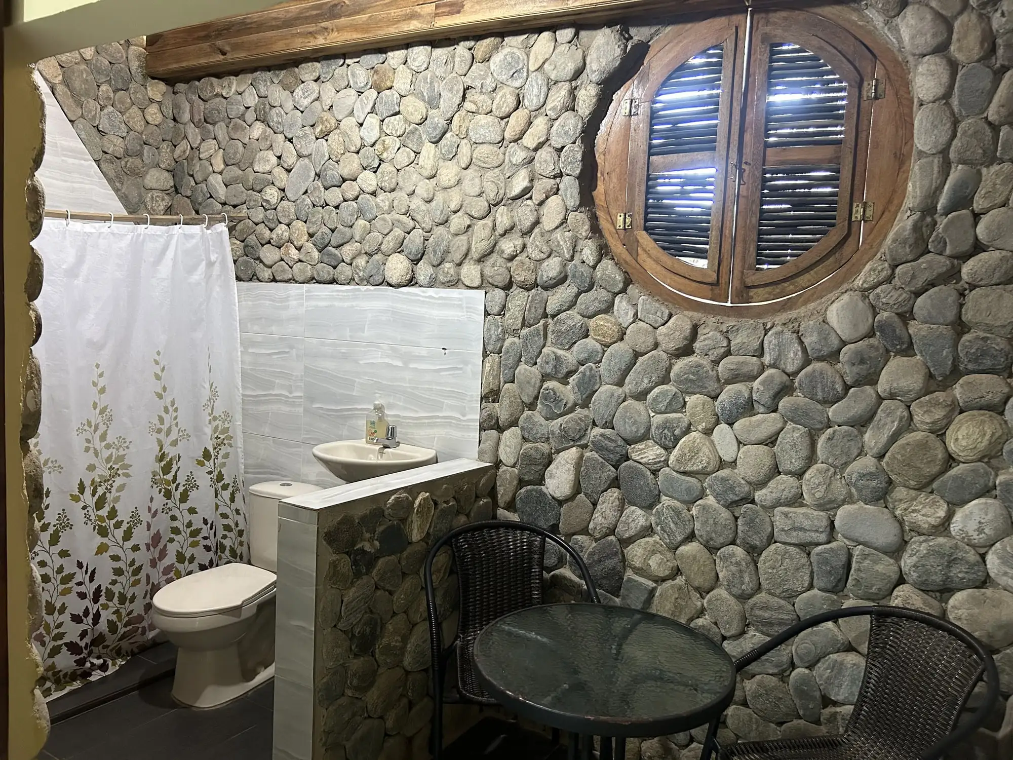 Baño privado con pared de piedra de río, ventana redonda con postigos, sanitario, lavamanos y ducha con cortina.