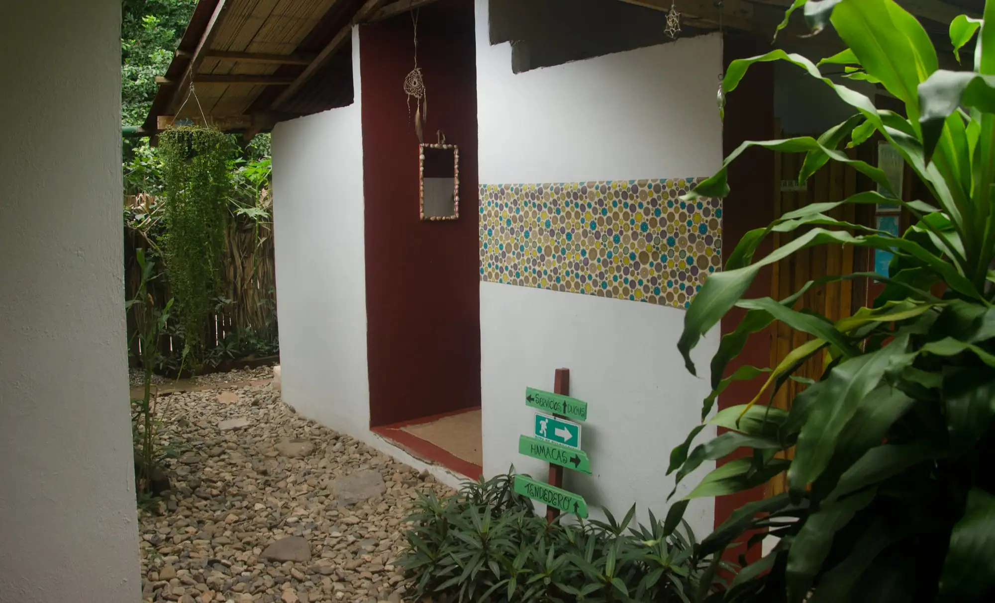Pasillo de grava hacia los baños compartidos, con una franja de mosaico y un letrero de madera pintado a mano que señala duchas y hamacas.