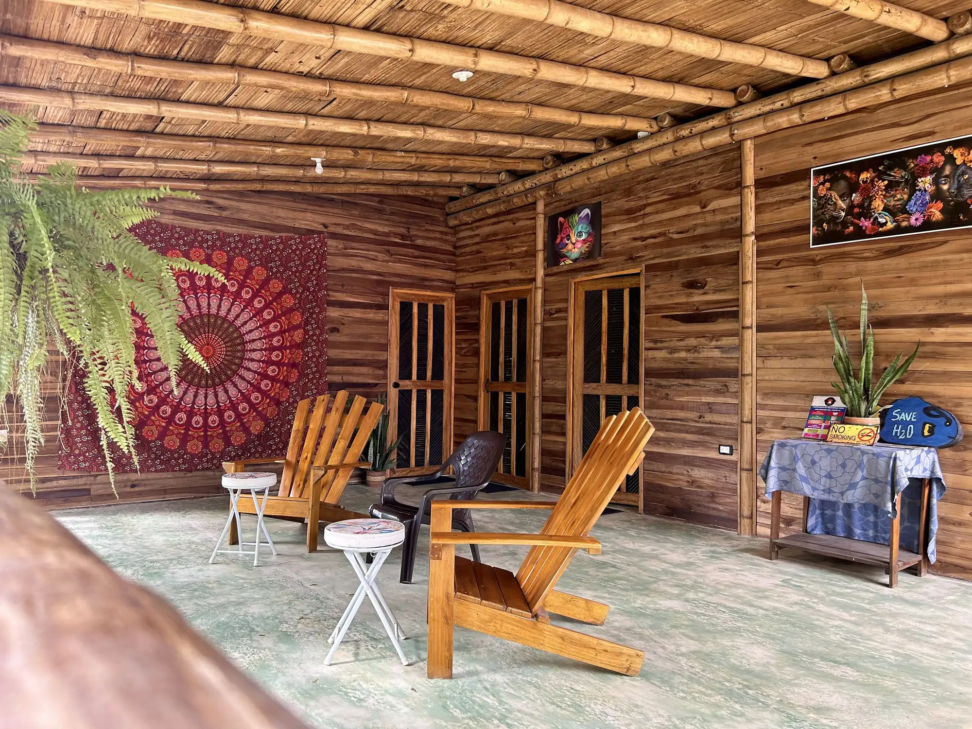 Terraza techada de madera frente a las habitaciones, con sillas de madera, un tapiz de mandala y helechos colgantes.
